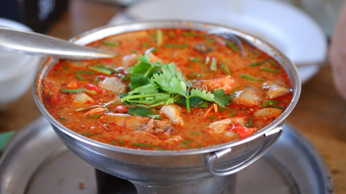 A steel pot of red, creamy tom yum soup with mushrooms and pieces of seafood, topped with cilantro, on a small burner stand