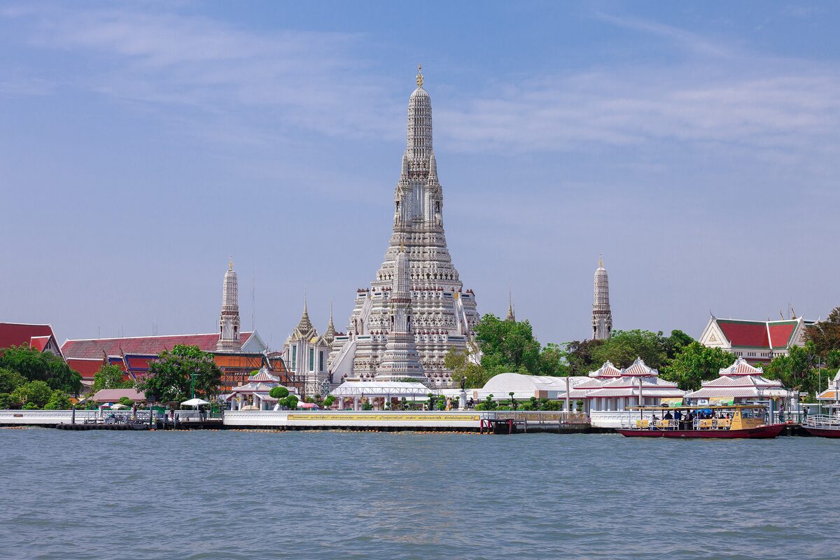 Wat Arun seen across the Chao Phraya River from the far bank, with its tall central prang and boats at the pier