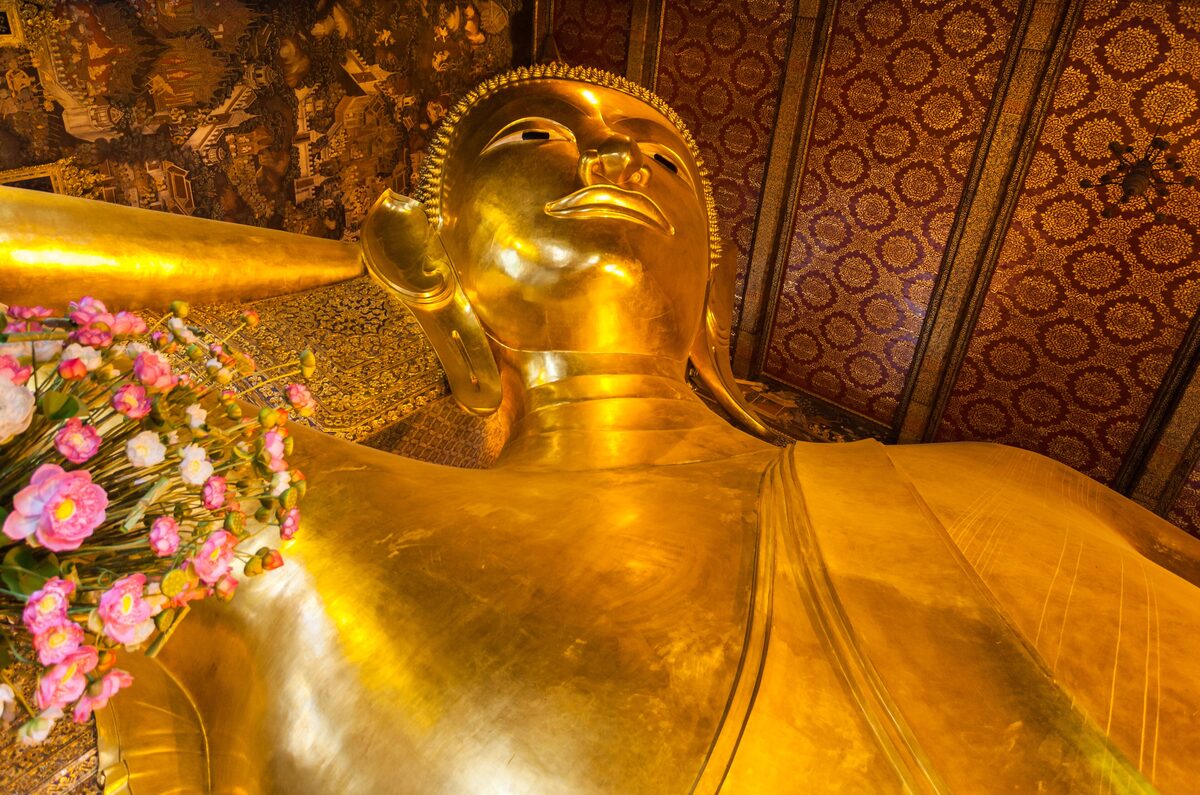 The gilded head and shoulder of the Reclining Buddha at Wat Pho under a patterned ceiling, with pink flower offerings at its side