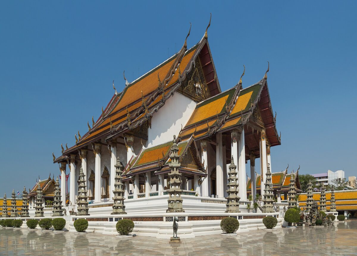 A white temple hall with gold and orange roofs and rows of small stupas in front, at Wat Suthat