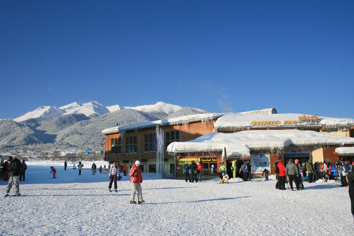 Skiers and walkers on the snow in front of the large timber-clad building at the Bansko gondola base, with mountains behind
