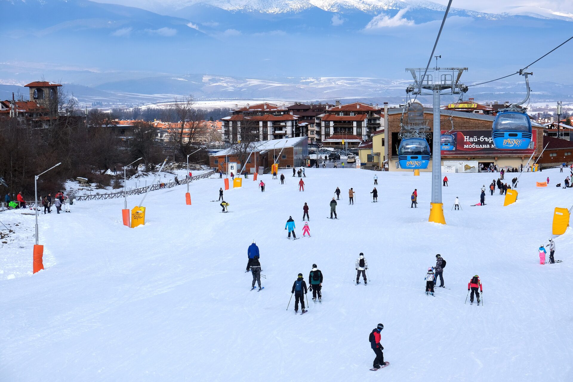Skiers on the snow below the Bansko gondola station, with cabins overhead and the town behind