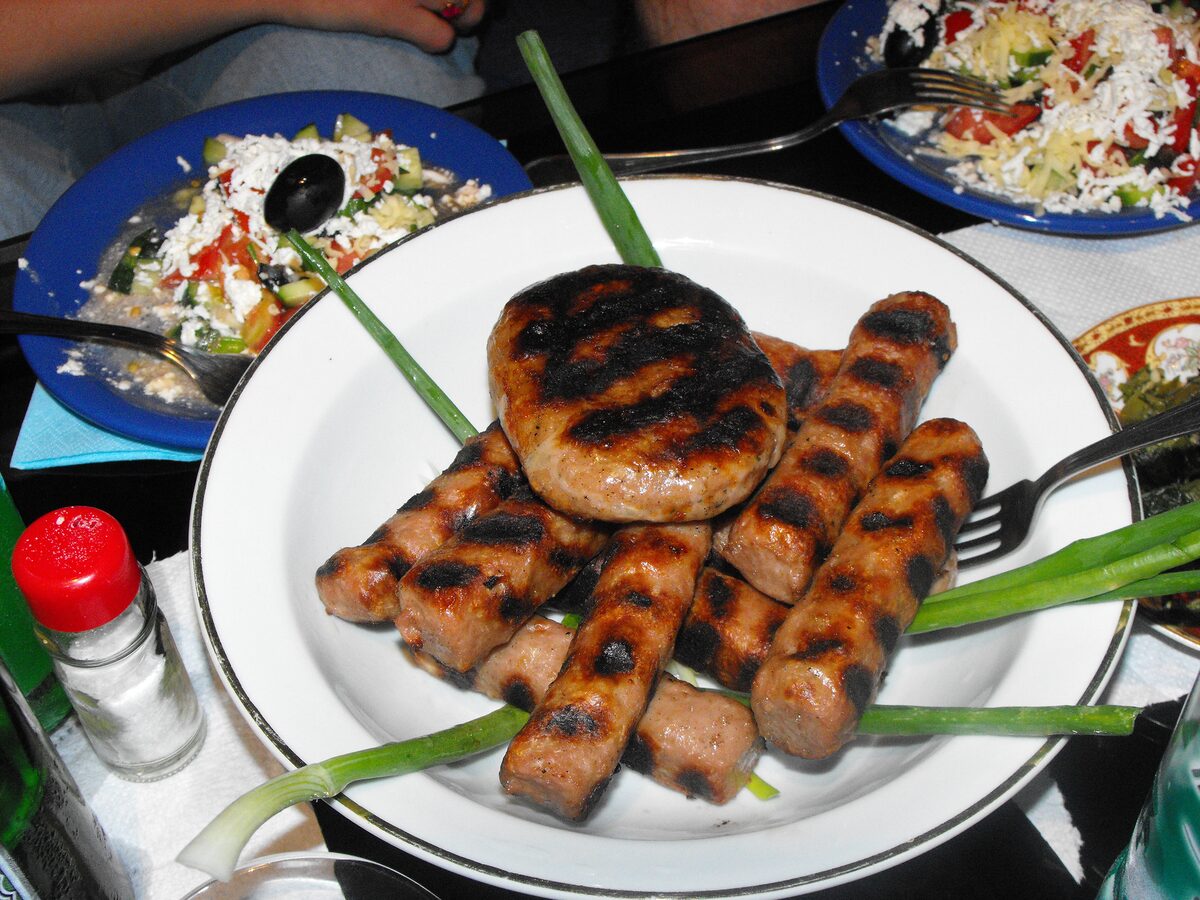 Grilled kebapche sausages and a round grilled patty on a white plate, with green onion and a salad behind