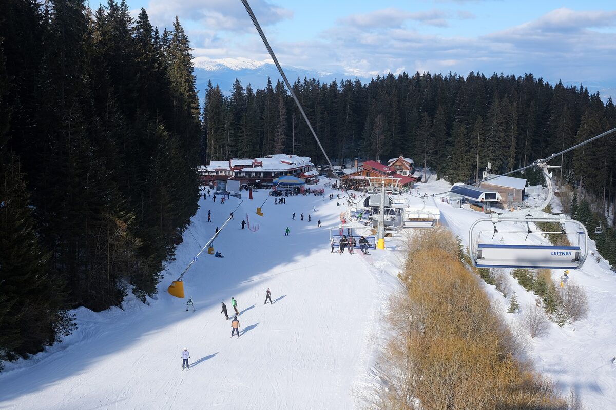 Skiers on a groomed piste beside the Todorka chairlift, with a lift station and huts at the bottom and pine forest alongside