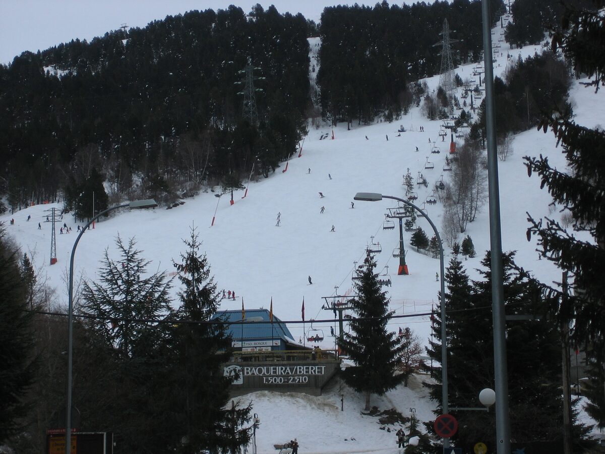 The Baqueira 1500 base in snow, with the Baqueira-Beret sign, a chairlift and pistes above the pines