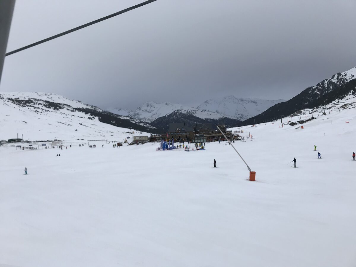 The Beret base area under grey cloud: a wooden lodge, a beginners' zone with skiers and a snow gun on the open slope