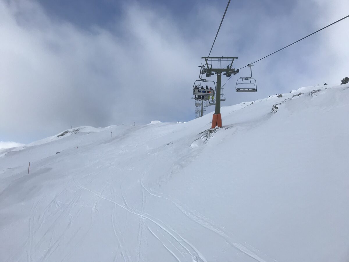 A chairlift climbing a groomed piste through fresh snow with cloud over the ridge