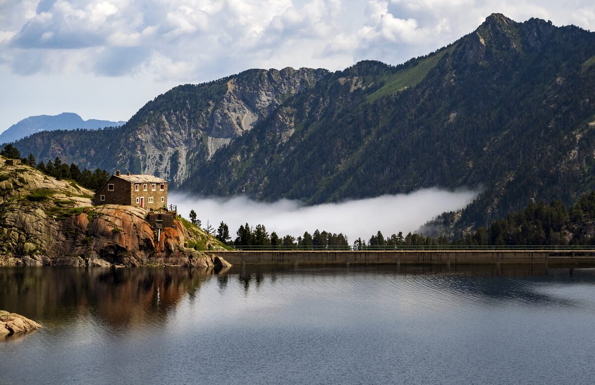The dam and stone refuge of the Colomèrs lake, with mountains and cloud behind