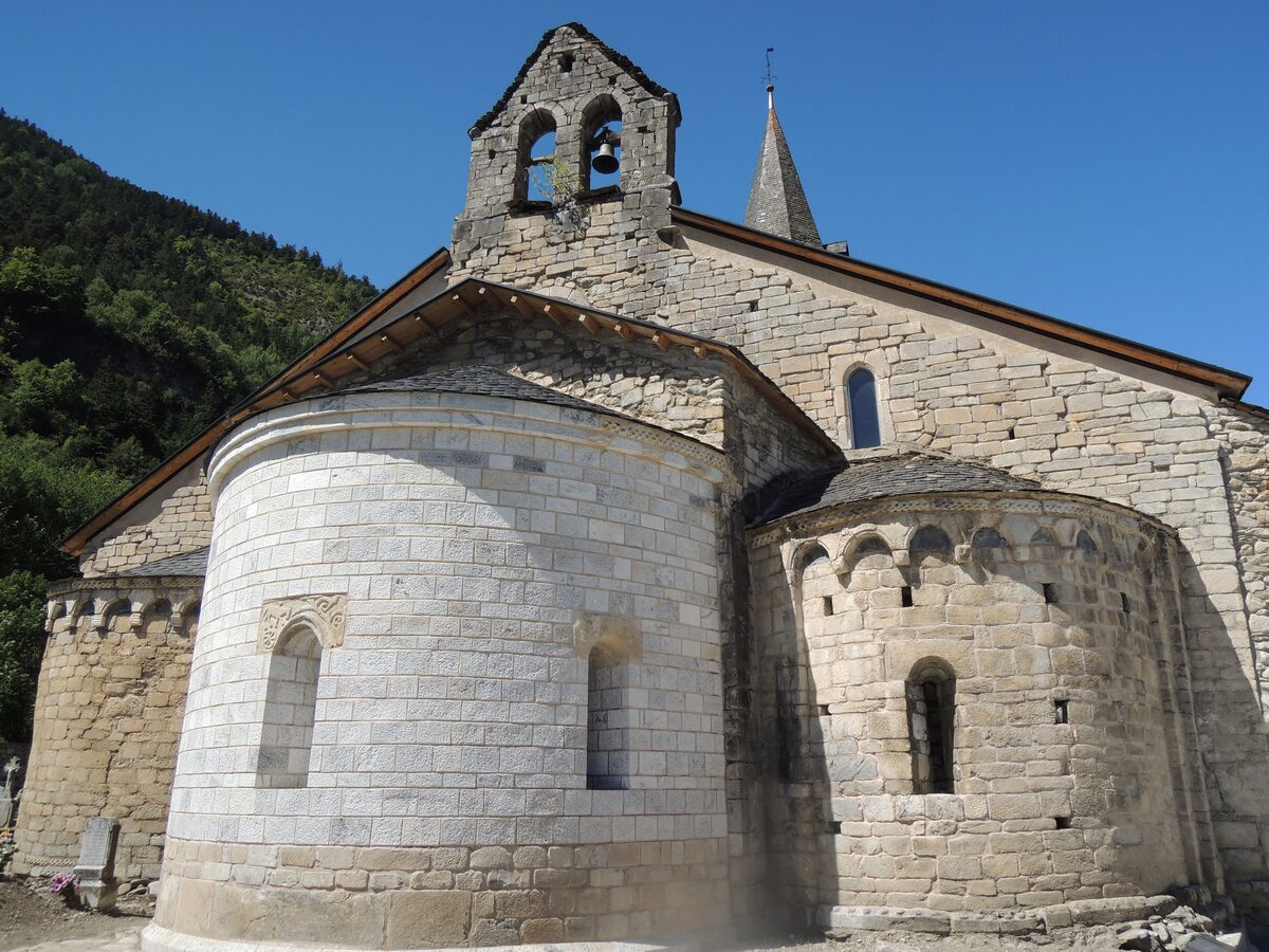 The apse and bell gable of Santa Maria d'Arties, a stone Romanesque church under a blue sky