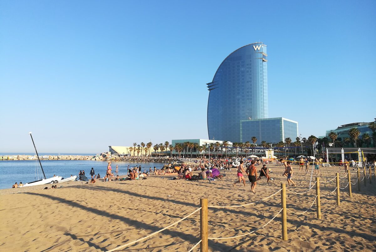 Sunny sand beach in Barceloneta with swimmers and the curved W hotel behind the palm-lined promenade