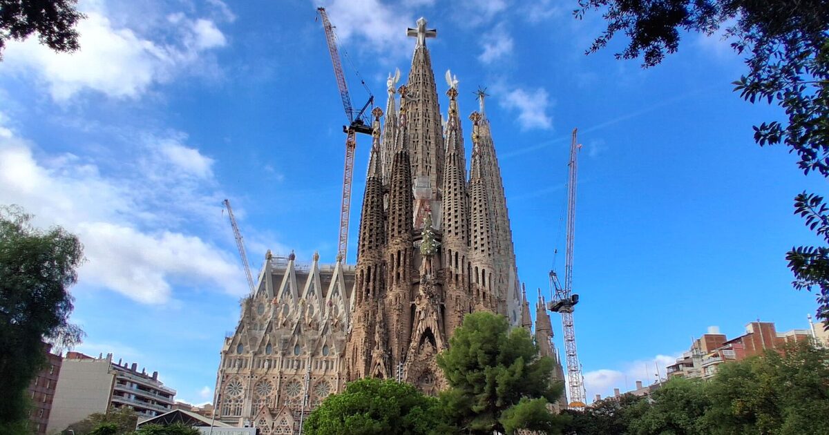 The Sagrada Família's Nativity facade and the central Tower of Jesus Christ with its cross, seen across the pond of Plaça de Gaudí under a blue sky