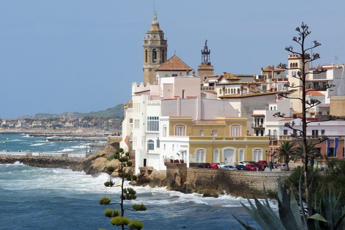 The Sant Bartomeu church tower above white houses on the Sitges seafront, with waves below