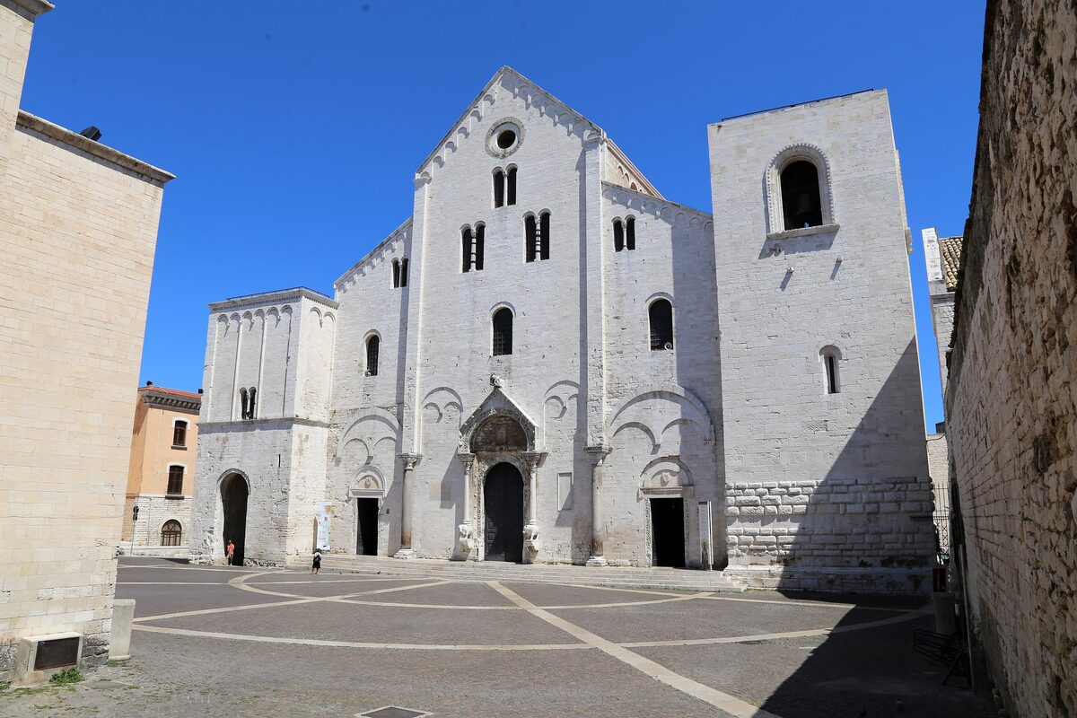 The white Romanesque facade of the Basilica of San Nicola on its paved square, with the central portal and the round window above