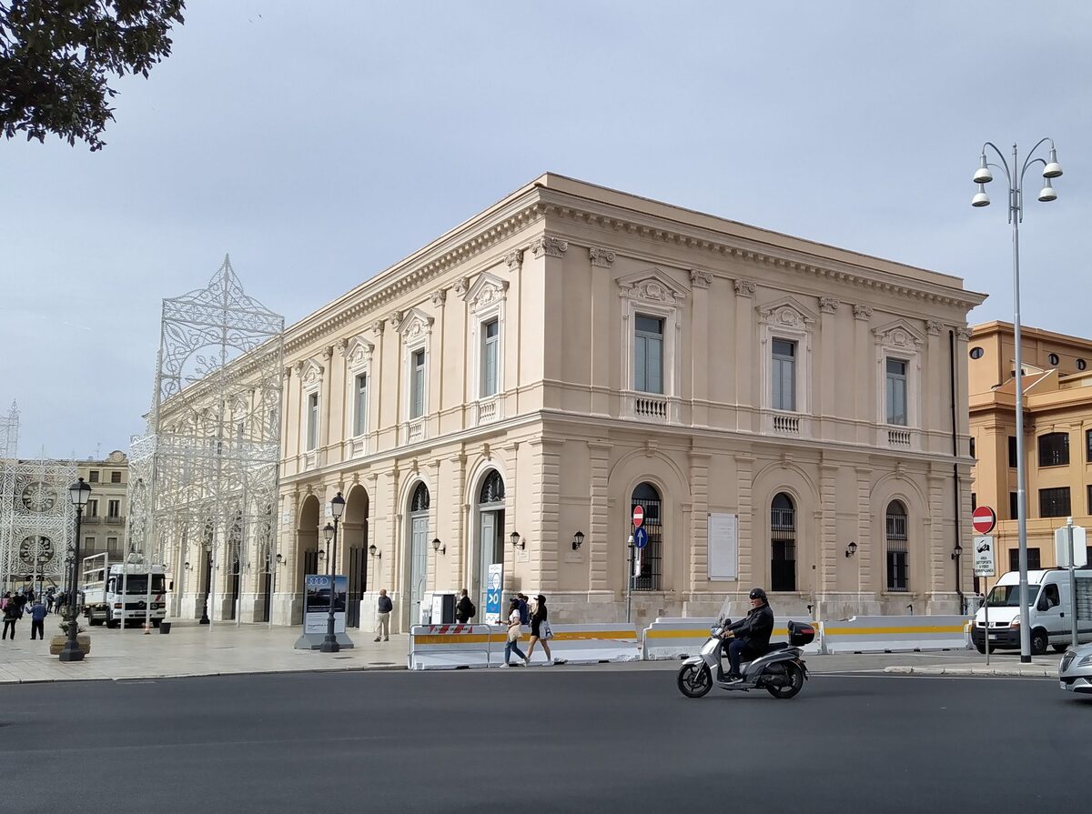 The former fish market building on Piazza del Ferrarese, a pale stone block with tall windows and a motorbike passing in the square