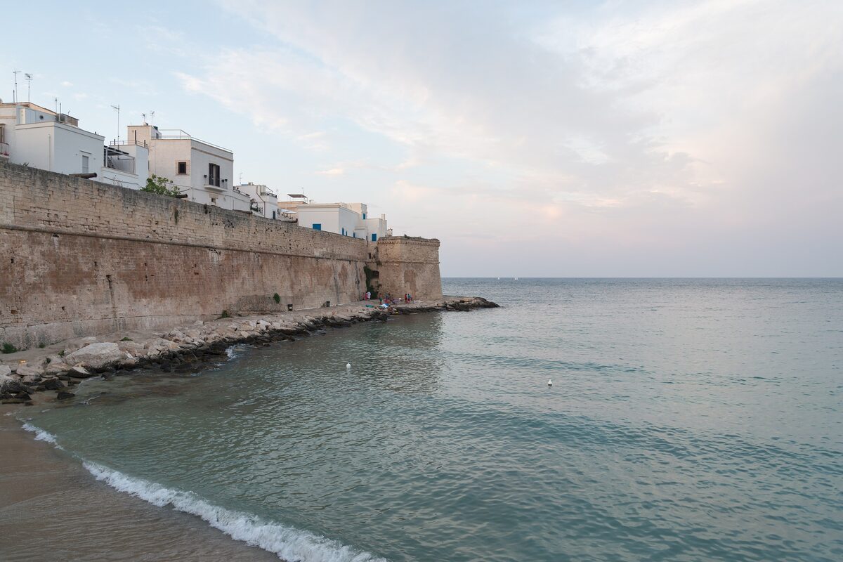 The old town wall of Monopoli above a small sandy cove, with calm sea at dusk