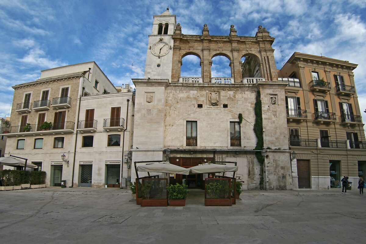 The clock tower of the Palazzo del Sedile over Piazza Mercantile, with tables set outside under umbrellas
