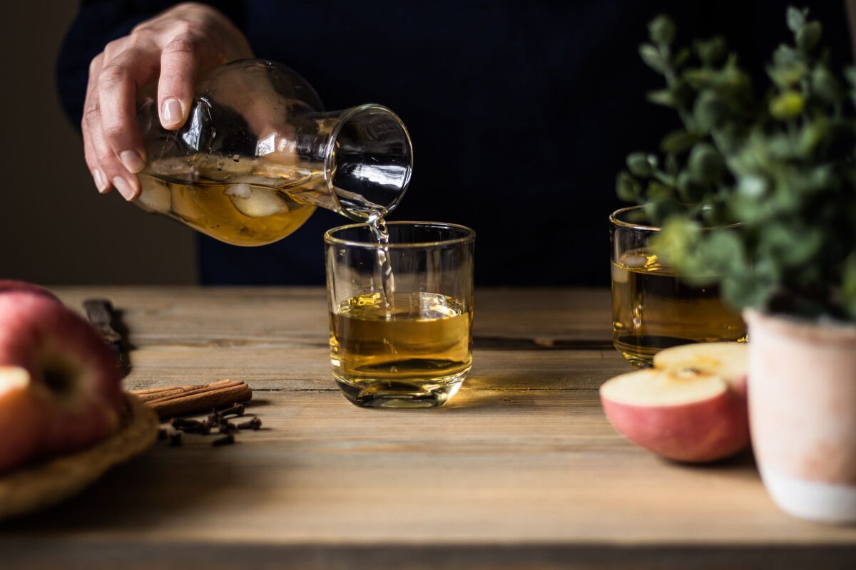 Golden cider being poured from a glass jug into tumblers, with red apples on a wooden table