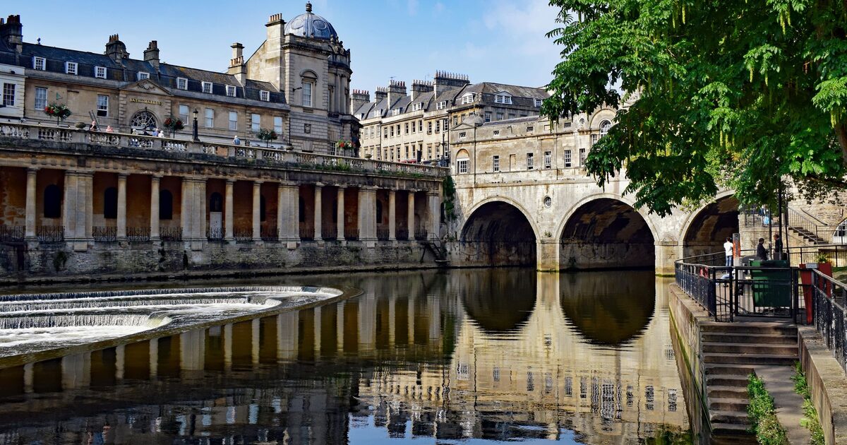 Pulteney Bridge and its arches reflected in the still River Avon in Bath, with a row of columns along the riverside and the weir on the left