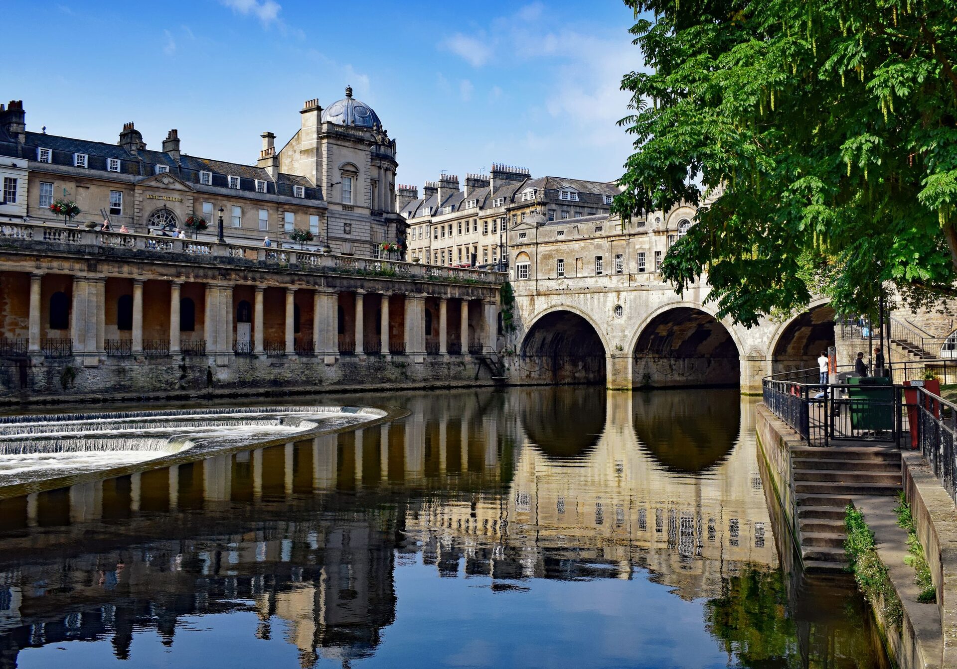 Pulteney Bridge and its arches reflected in the still River Avon in Bath, with a row of columns along the riverside and the weir on the left
