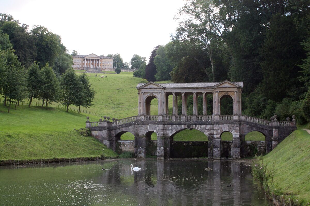 The Palladian Bridge at Prior Park, a covered bridge with columns over a lake, among trees, with Bath in the distance