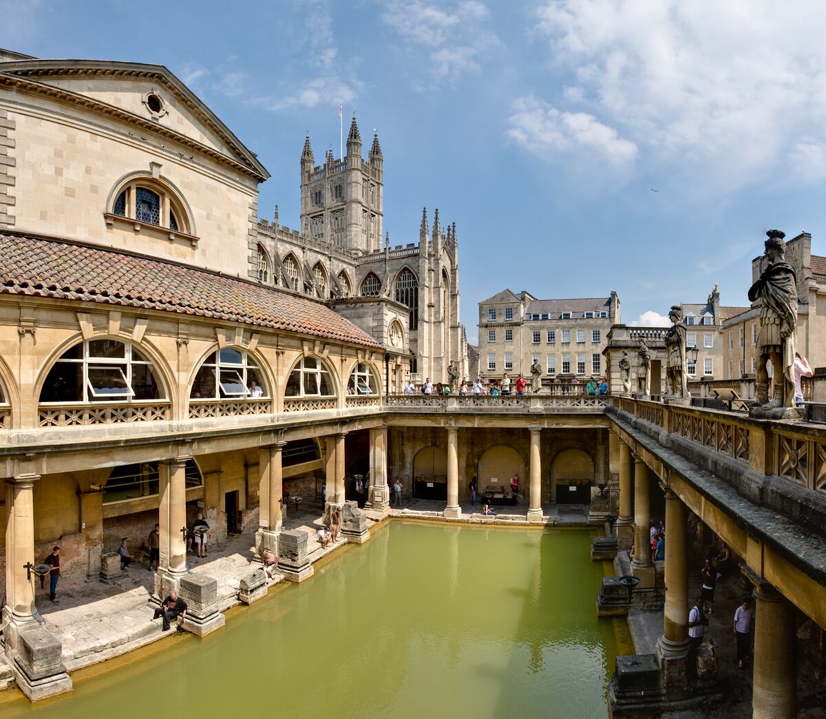 The green water of the Great Bath at the Roman Baths, with stone columns around it and the Abbey tower behind