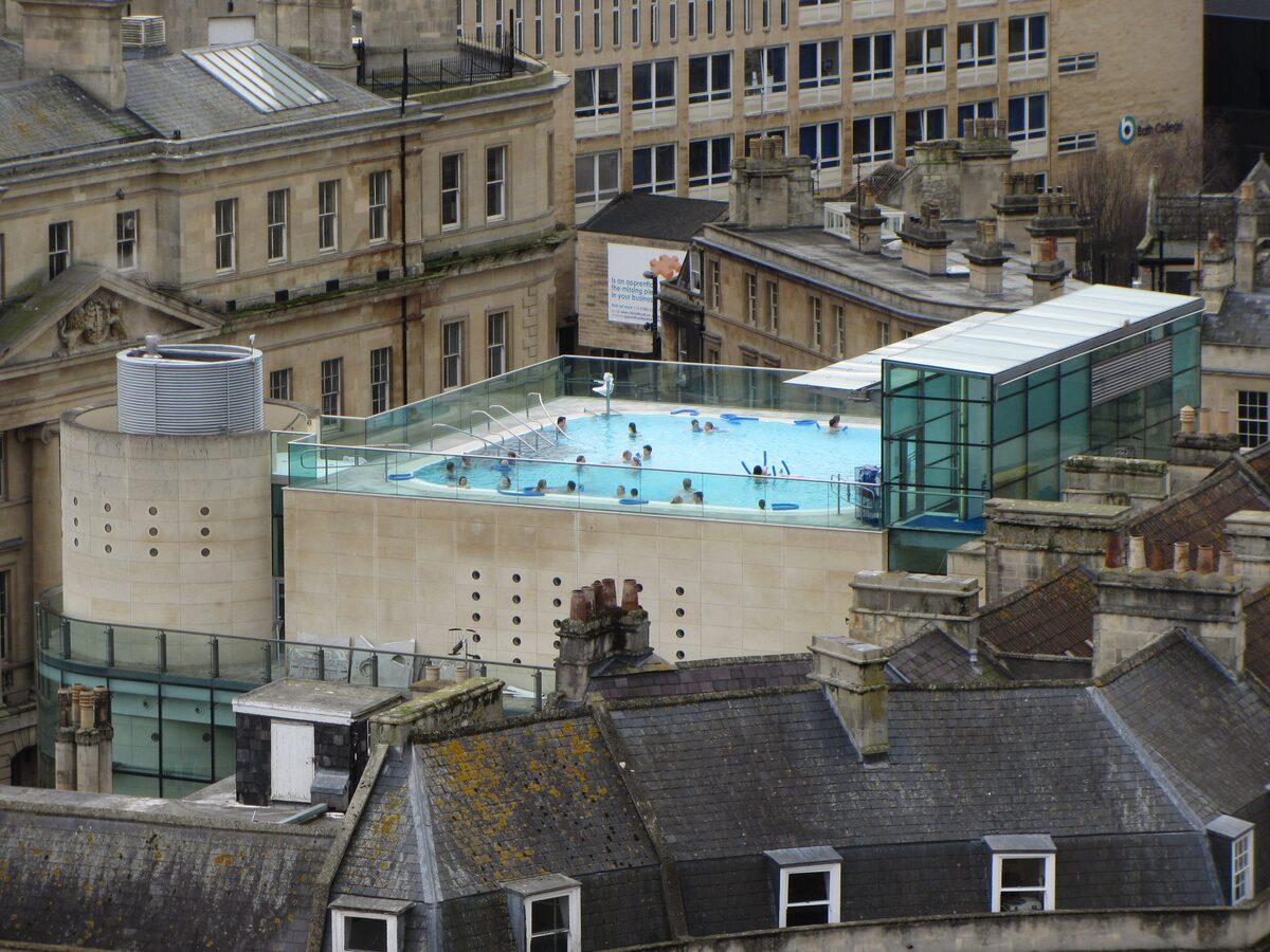 The rooftop pool of Thermae Bath Spa, full of bathers, seen from above among the stone roofs of Bath