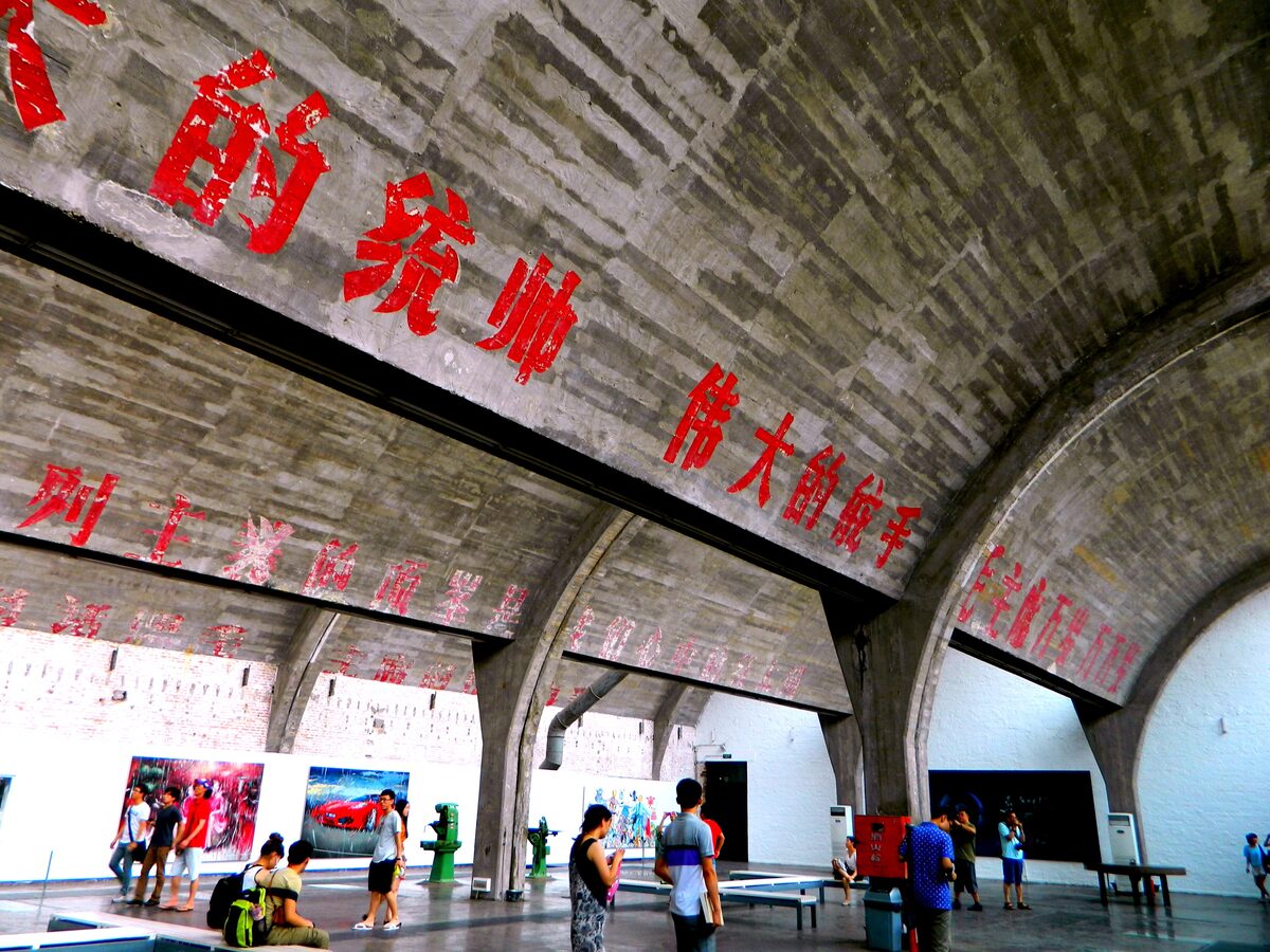Visitors under the concrete vaults of a former factory hall in the 798 Art Zone, now an exhibition space, with red slogans from the factory era painted on the arches