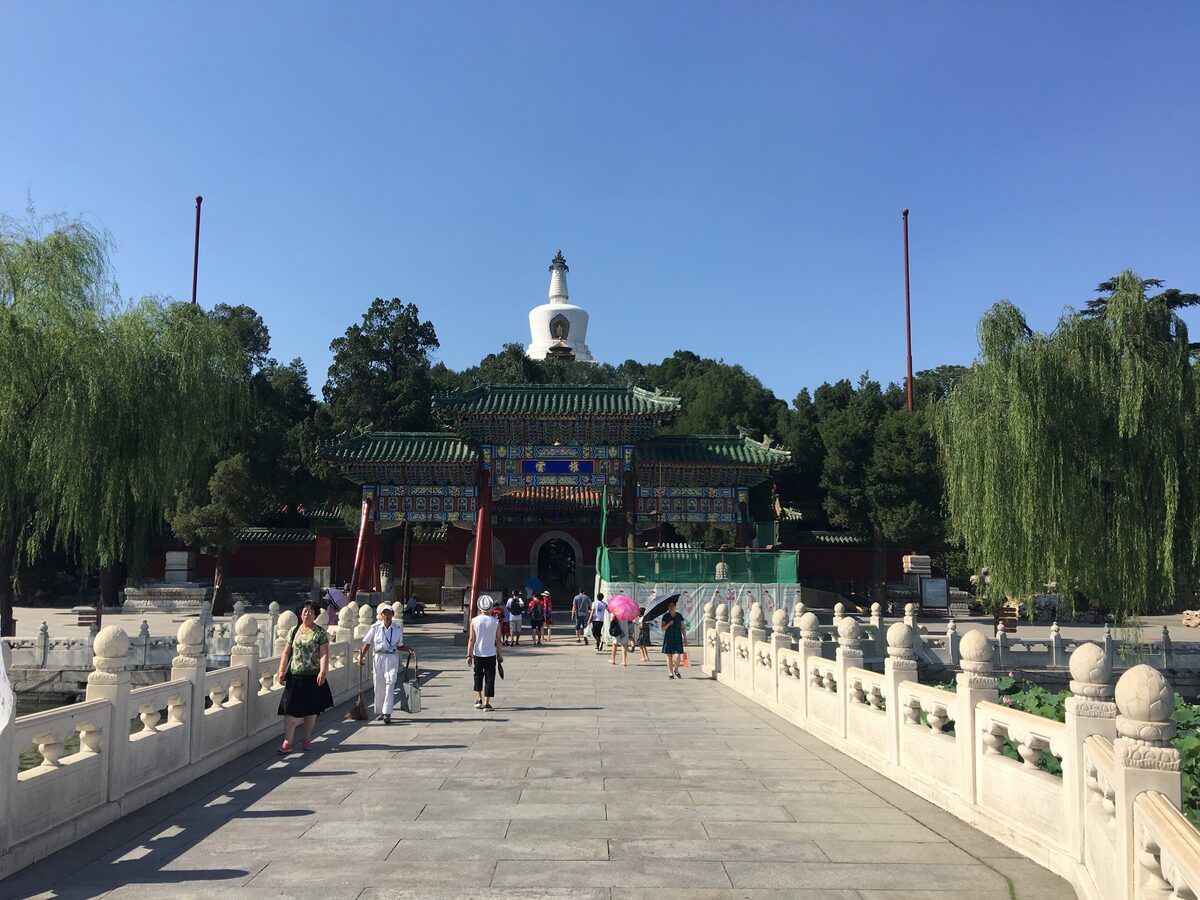 A white marble bridge in Beihai Park leading to a painted gate, with the white dagoba of Jade Flower Island above the trees and willows on both sides
