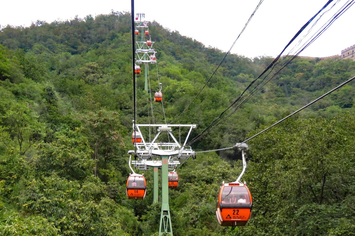 Orange cable car cabins on their cables climbing the green wooded slope at Mutianyu, with a stretch of wall on the ridge at top right