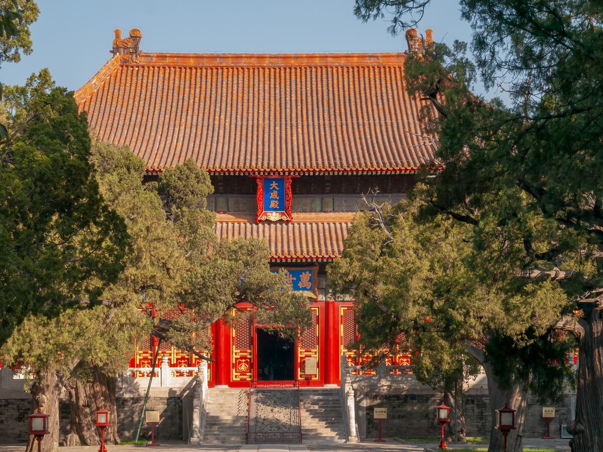 A hall of the Confucius Temple with an orange-tiled roof and red doors, framed by old cypress trees