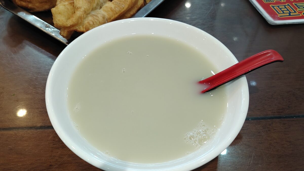 A white bowl of grayish-white douzhi with a red spoon, and fried dough at the edge of the table