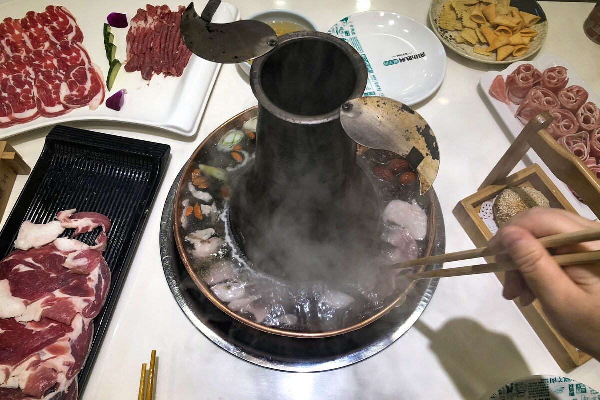 A copper charcoal hot pot steaming on a table, with plates of thinly sliced lamb and a hand dipping meat with chopsticks