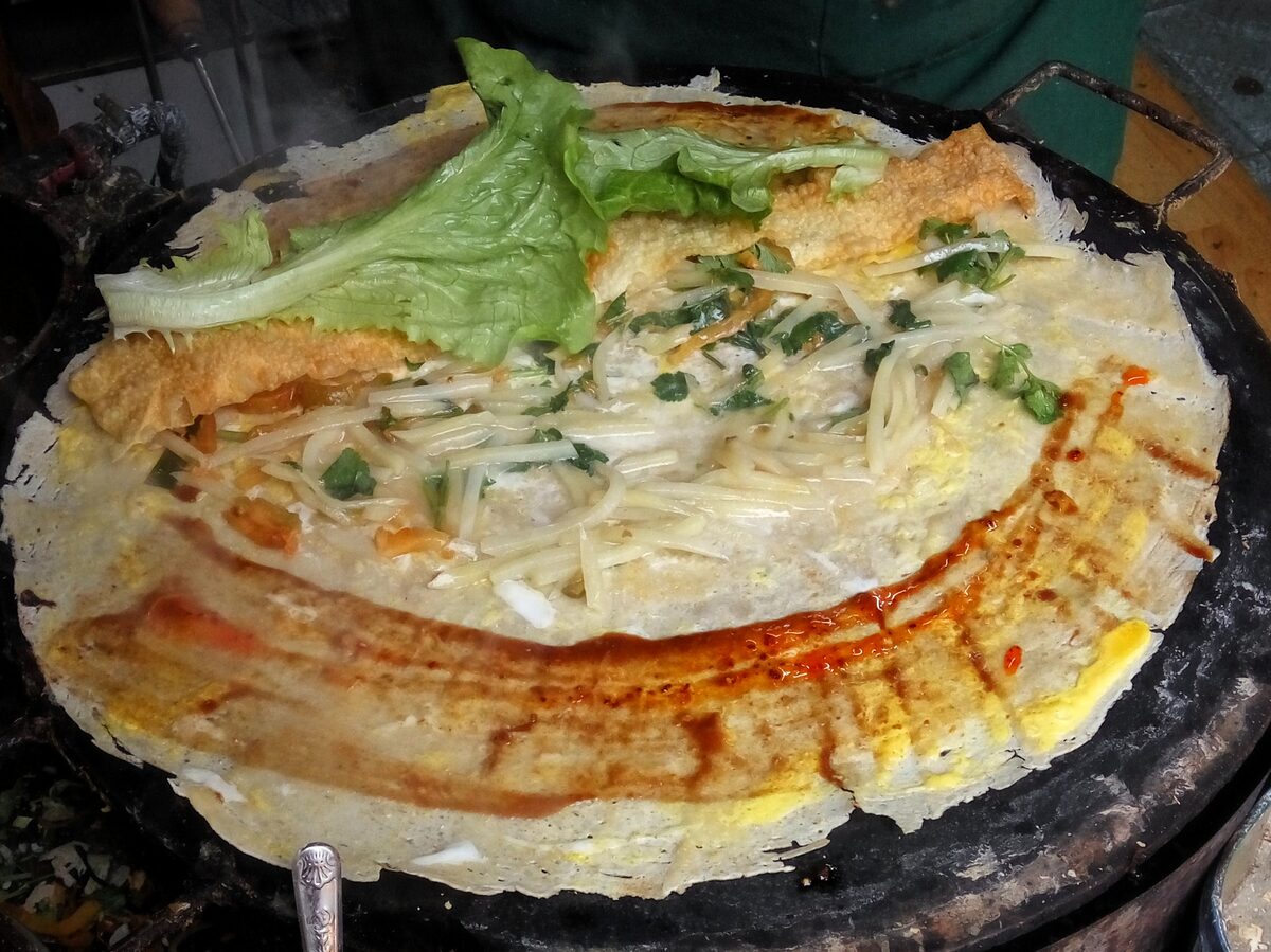 A jianbing crêpe on a round griddle, topped with egg, lettuce, scallion, cilantro, a crisp cracker and a streak of red chili sauce