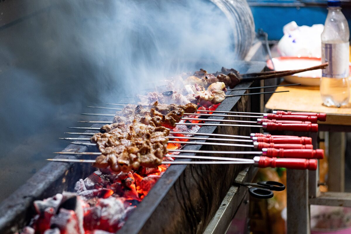 Rows of lamb skewers grilling over glowing charcoal in a long trough, with smoke rising