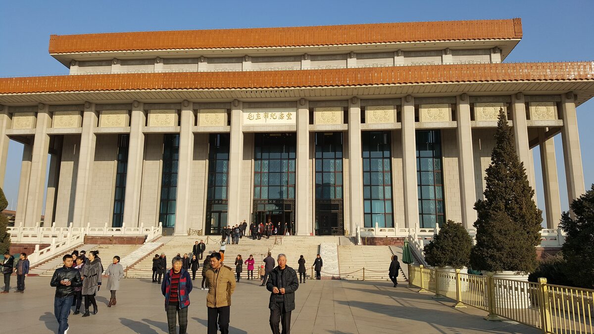 The Chairman Mao Memorial Hall by day, a wide colonnaded building with an orange-tiled double roof, visitors on its forecourt and a blue sky above