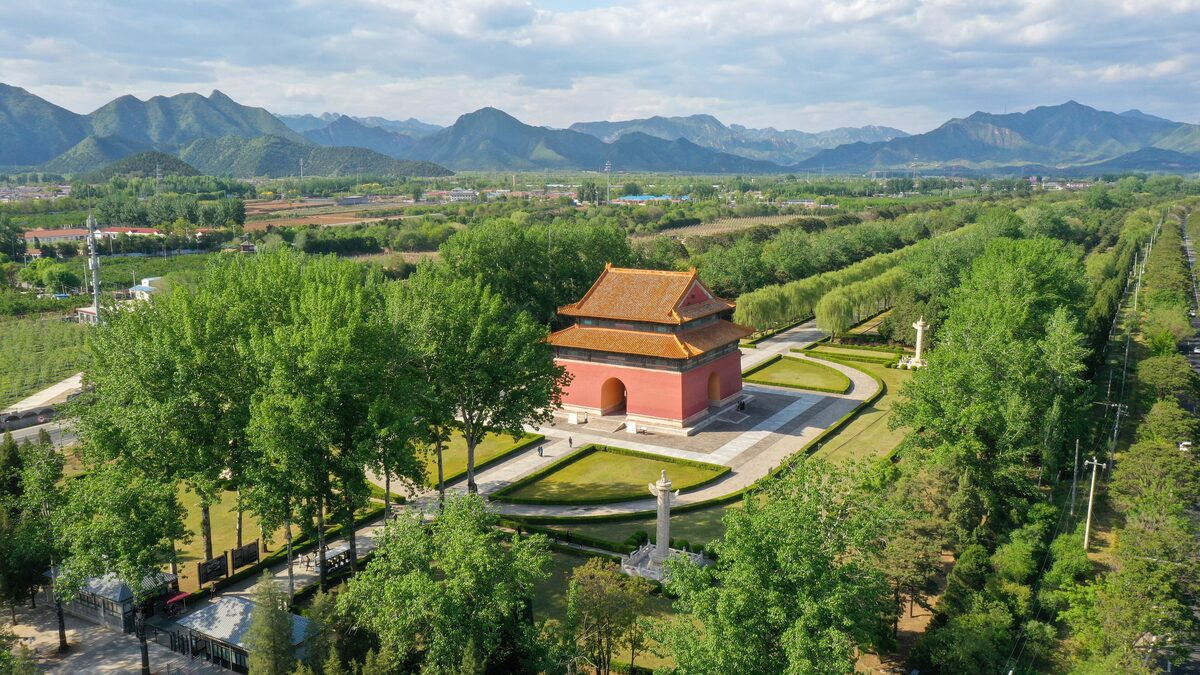 A red pavilion at the Ming Tombs seen from above, surrounded by tree-lined paths with mountains behind