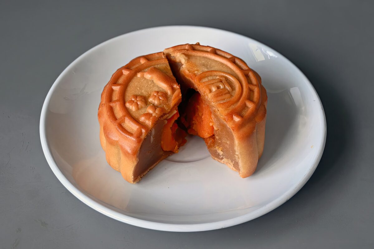 A mooncake cut open on a white plate, showing lotus seed paste and a salted egg yolk inside its golden-brown pressed crust