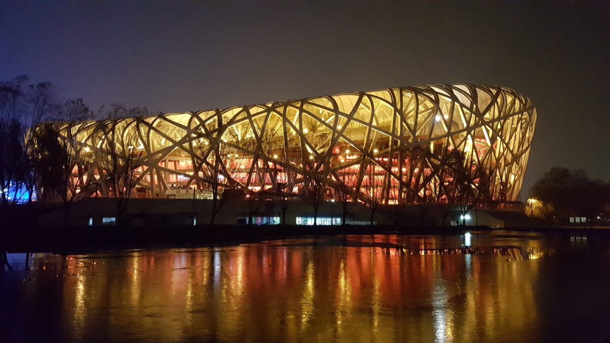 The Bird's Nest National Stadium lit up at night, its steel lattice glowing and reflected in the water in front