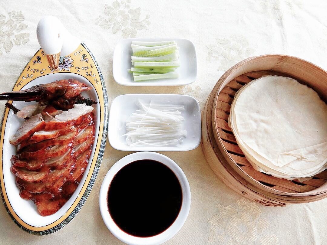 Sliced roast Peking duck on a platter with a bamboo basket of thin pancakes, cucumber, scallion and dark sweet bean sauce