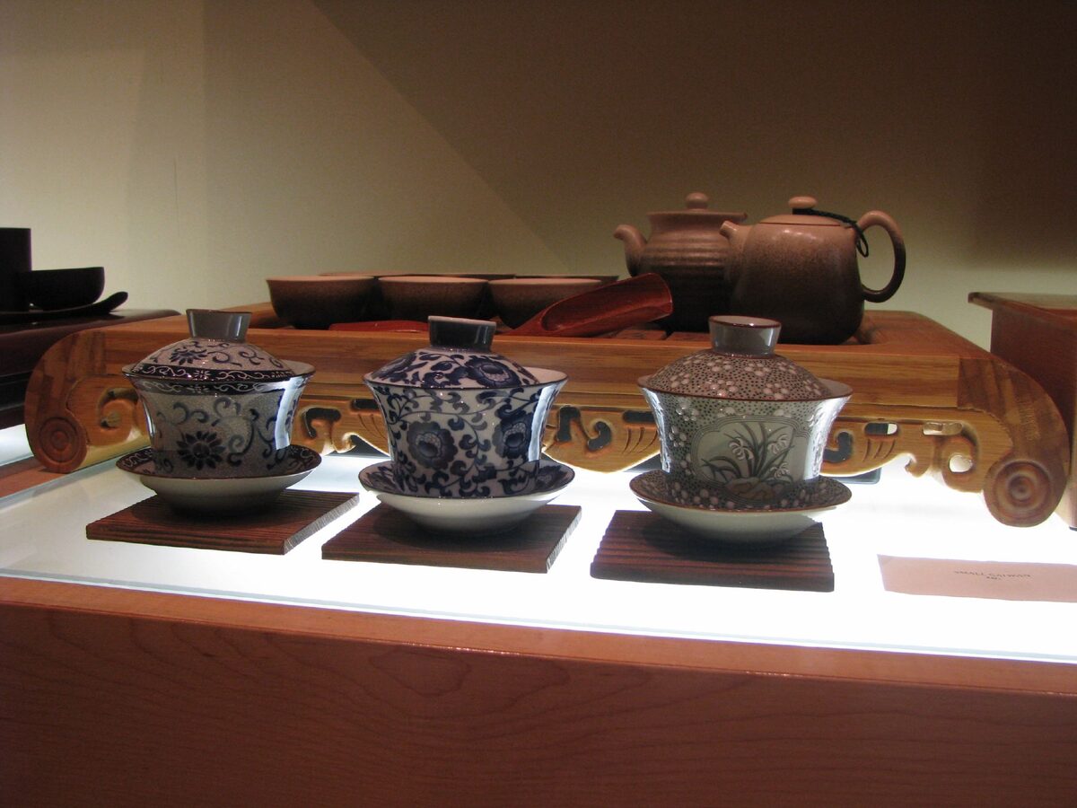 Three lidded porcelain gaiwan tea cups on saucers on a wooden tea table, with clay teapots behind