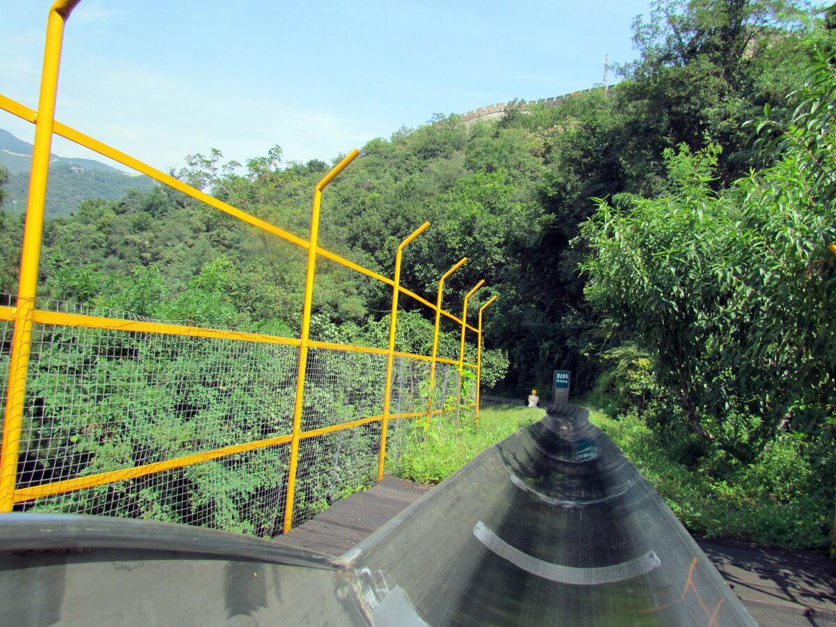 The metal toboggan track at Mutianyu, seen from the sled, curving through green trees beside a yellow safety fence