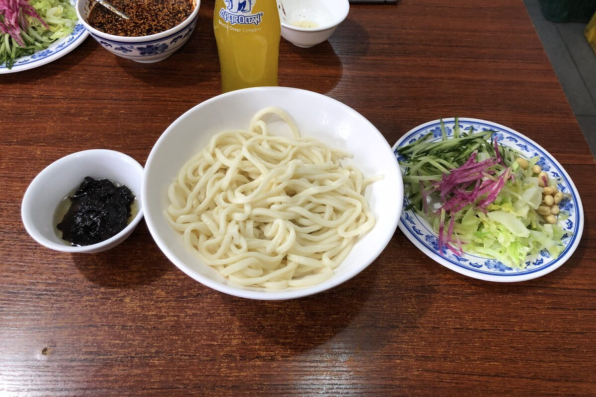 A bowl of plain wheat noodles with a dish of dark soybean sauce and a plate of shredded vegetables beside it