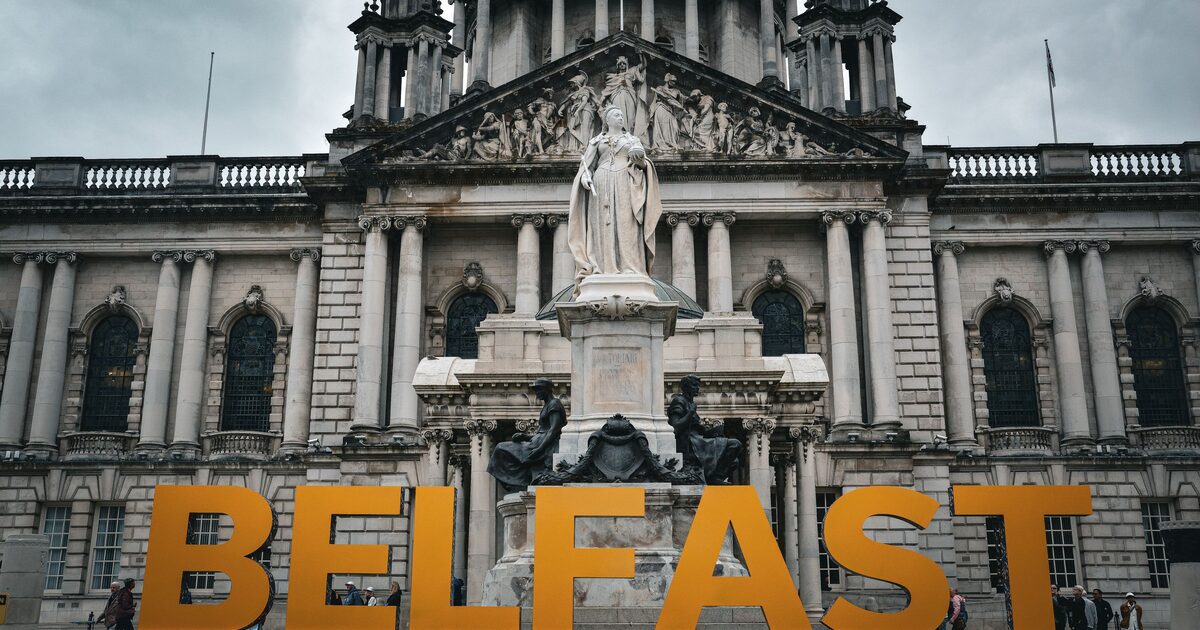 Belfast City Hall with its white stone front, green dome and statue of Queen Victoria, behind big orange letters spelling BELFAST