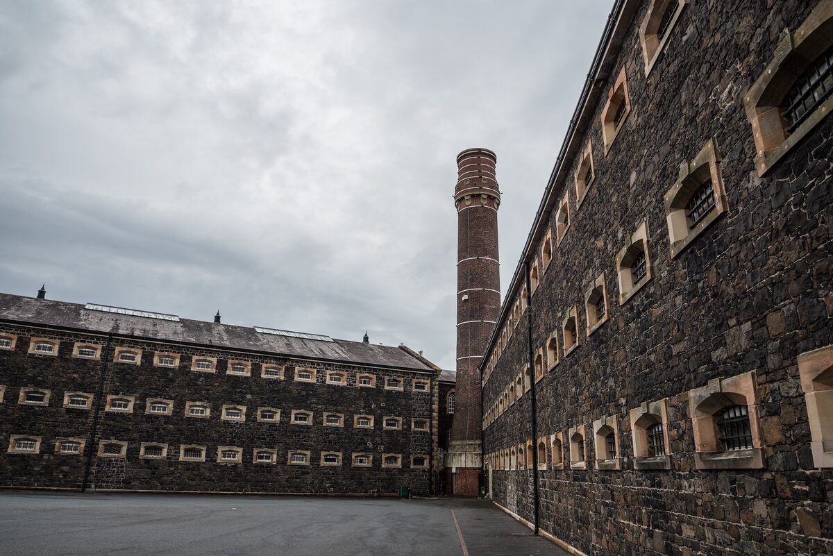 The long black basalt wings of Crumlin Road Gaol with rows of small barred windows and a tall chimney between them