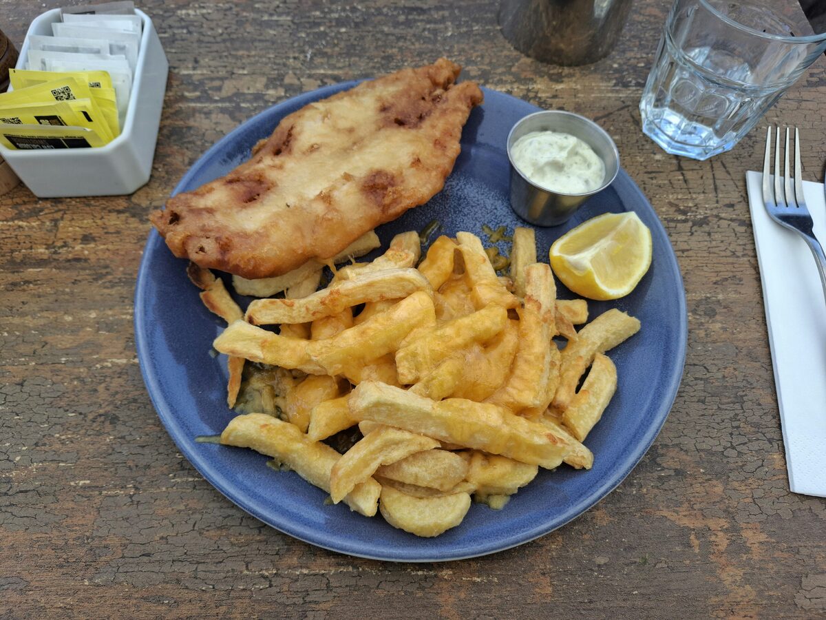 A piece of battered fish with thick chips, lemon and tartare sauce on a blue plate