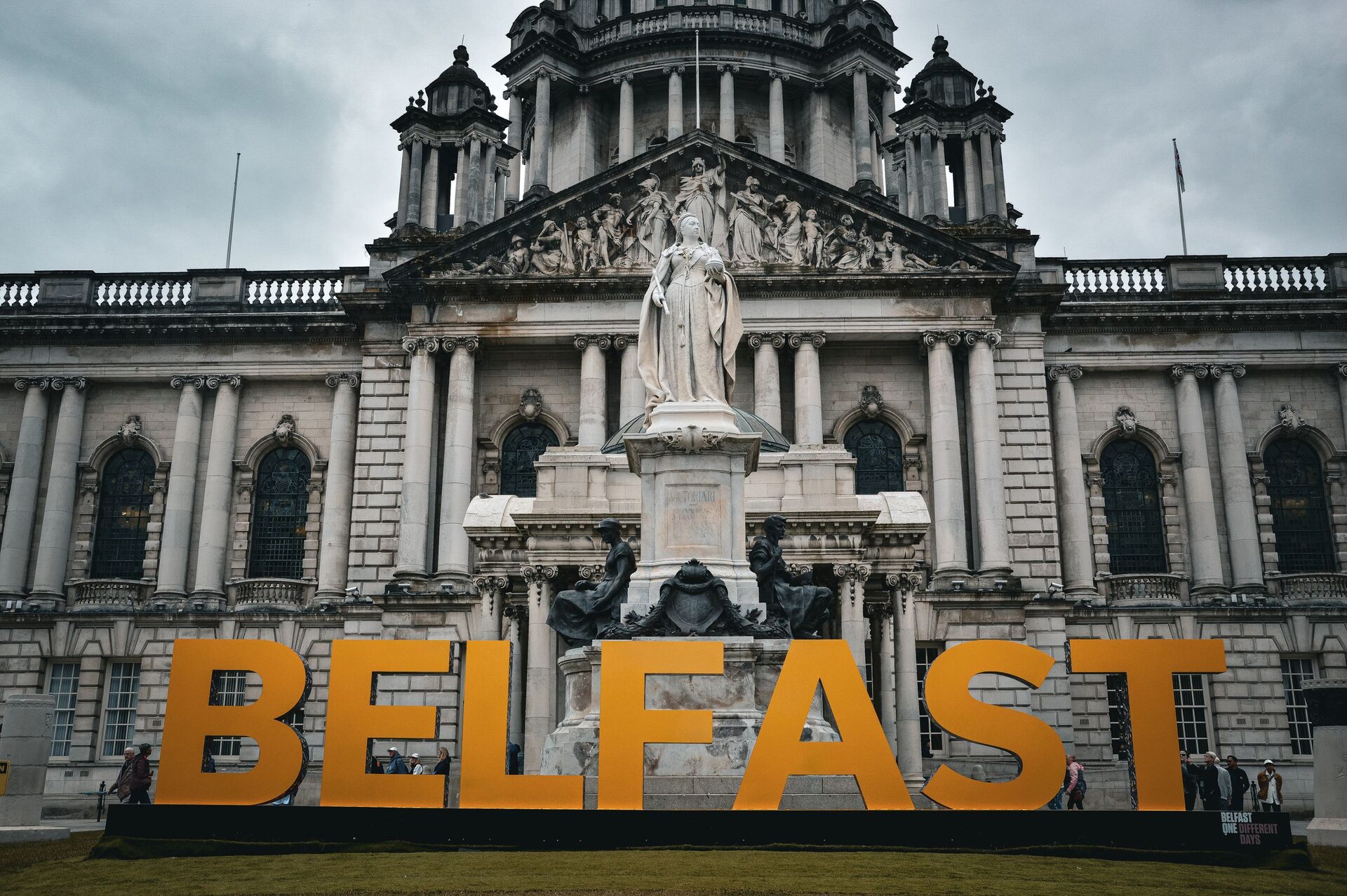 Belfast City Hall with its white stone front, green dome and statue of Queen Victoria, behind big orange letters spelling BELFAST