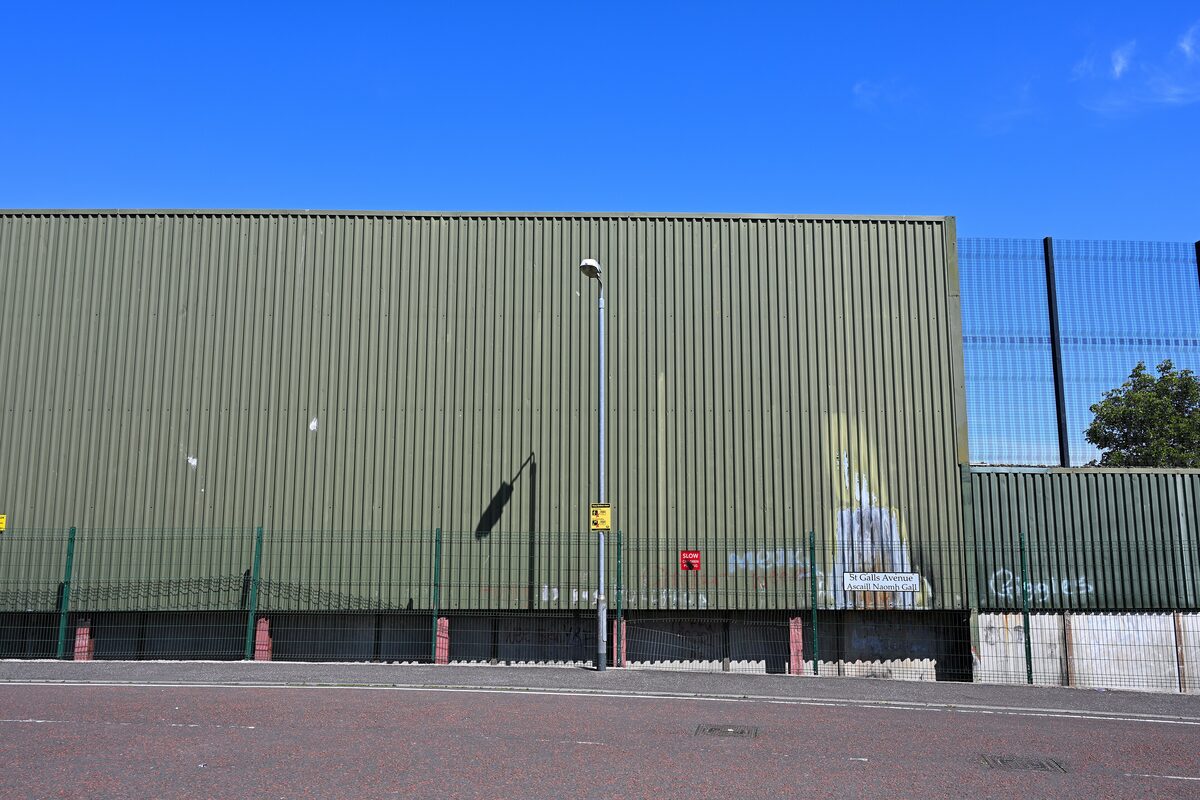 A tall green metal peace wall topped with fencing, running along a street in west Belfast under a blue sky