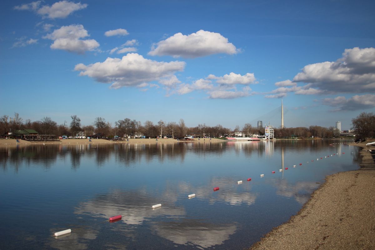 Calm water and the beach at Ada Ciganlija lake, with the pylon of the Ada Bridge in the distance