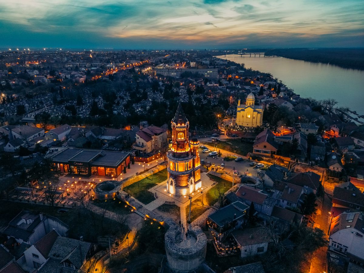 Gardoš Tower lit up at dusk above the rooftops of Zemun, with the Danube on the right
