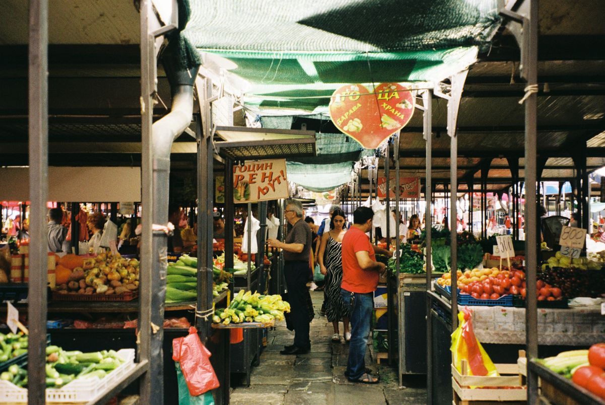 Vegetable stalls and shoppers under green shade nets at an open-air market in Belgrade